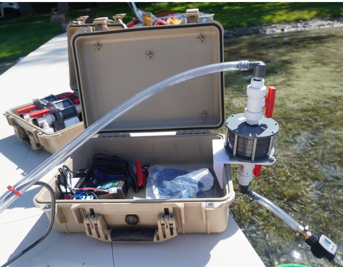 An open beige hard-shell carrying case on a dock, connected with clear tubing to a small pump or filter unit for eRNA surveying. Another similar case sits behind it, and water is visible in the background outdoors.
