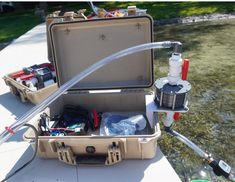 An open beige hard-shell carrying case on a dock, connected with clear tubing to a small pump or filter unit for eRNA surveying. Another similar case sits behind it, and water is visible in the background outdoors.
