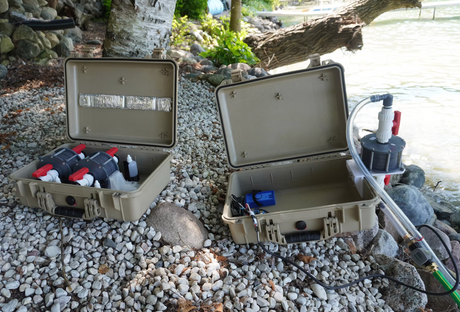 Two beige waterproof hard cases open on a rocky lakeshore, containing electronic monitoring devices for eRNA surveying and connected tubing, with lake and trees in the background.