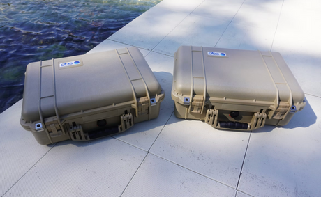 Two tan hard-shell protective cases with latches and handles sit on a light tiled surface next to a lake.