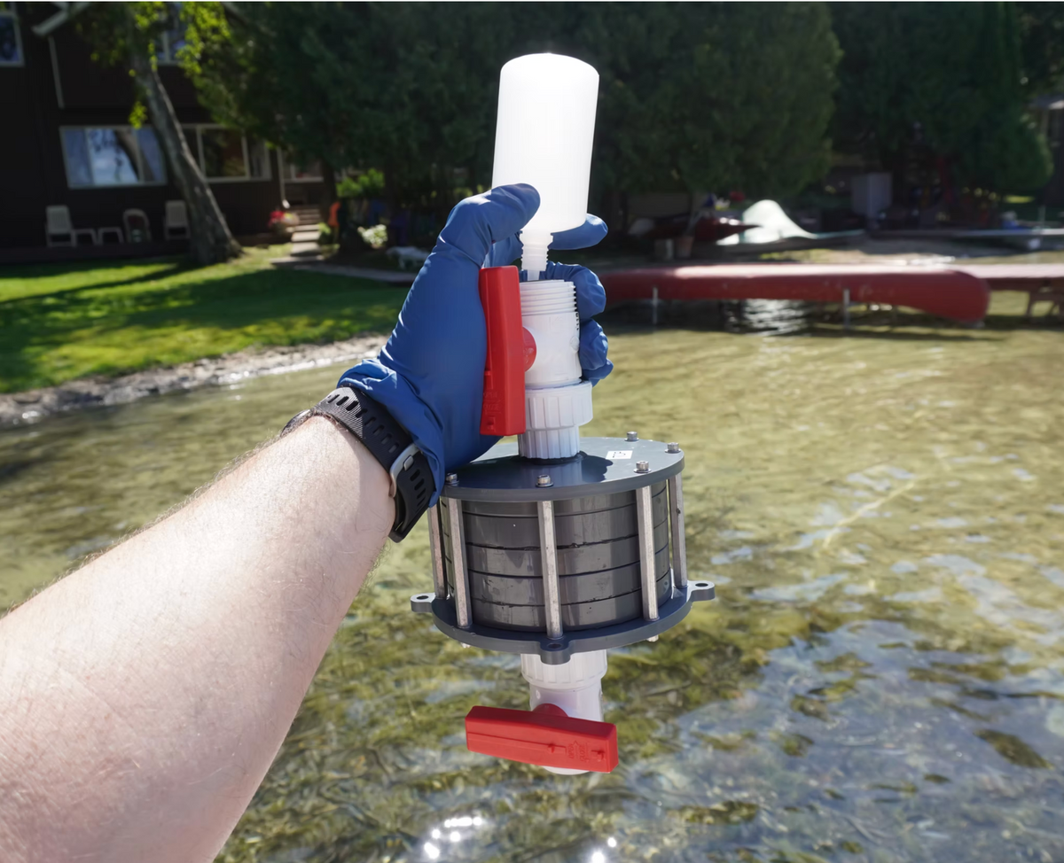 A person wearing a blue glove holds a manual water filter pitcher outdoors near a lake, with grass, trees, and a dock visible in the background.