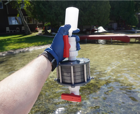A person wearing a blue glove holds a manual water filter pitcher outdoors near a lake, with grass, trees, and a dock visible in the background.
