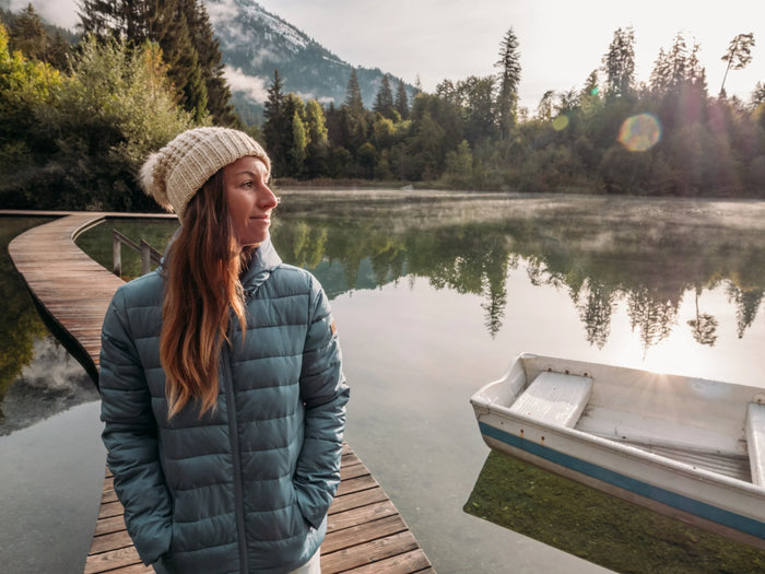 Woman in winter jacket and beanie stands on lakeside boardwalk with boat, mountain view, Lake Pulse